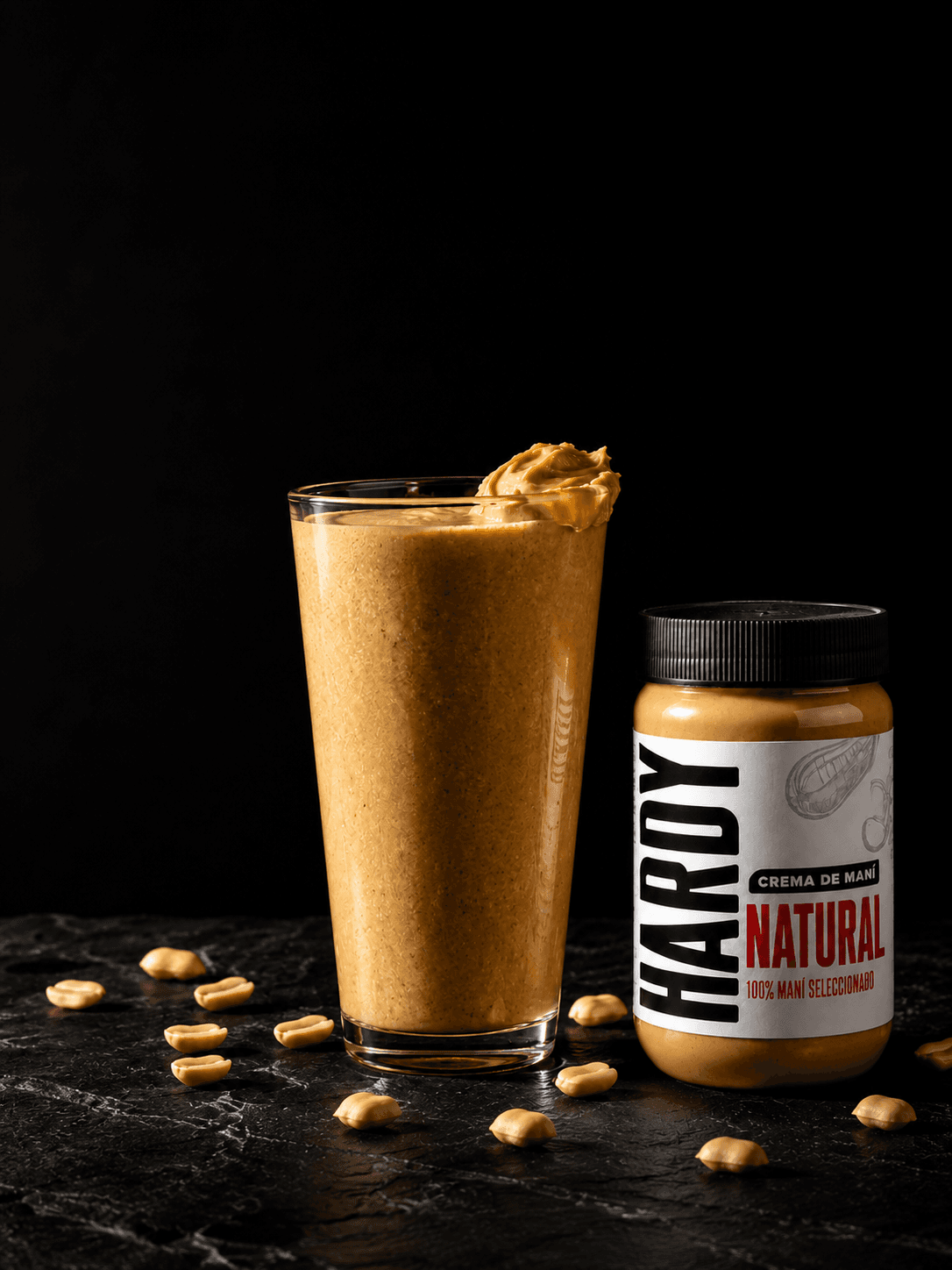 Smoothie de crema de maní Hardy en vaso alto con maníes sueltos sobre piedra negra